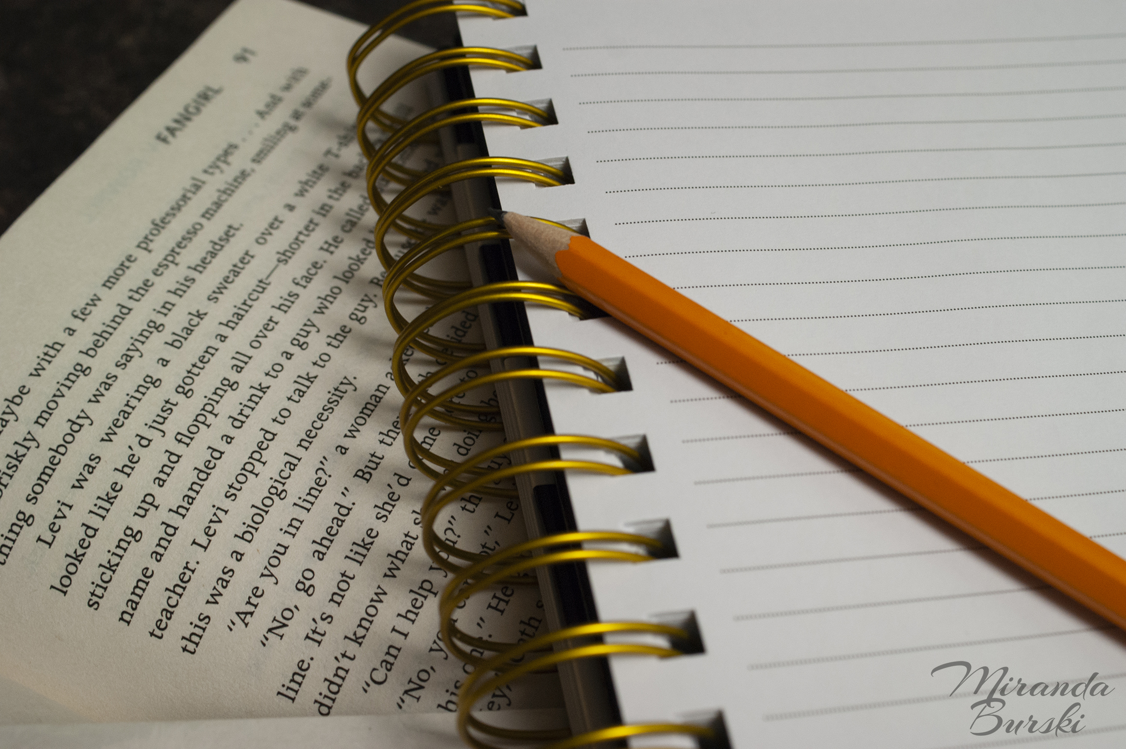 A pencil and notebook on top of an open copy of Fangirl, by Rainbow Rowell