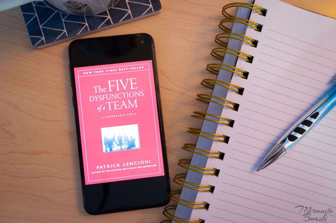 A digital copy of the cover of The Five Dysfunctions of a Team, by Patrick Lencioni, beside a notebook, pen and coffee mug.