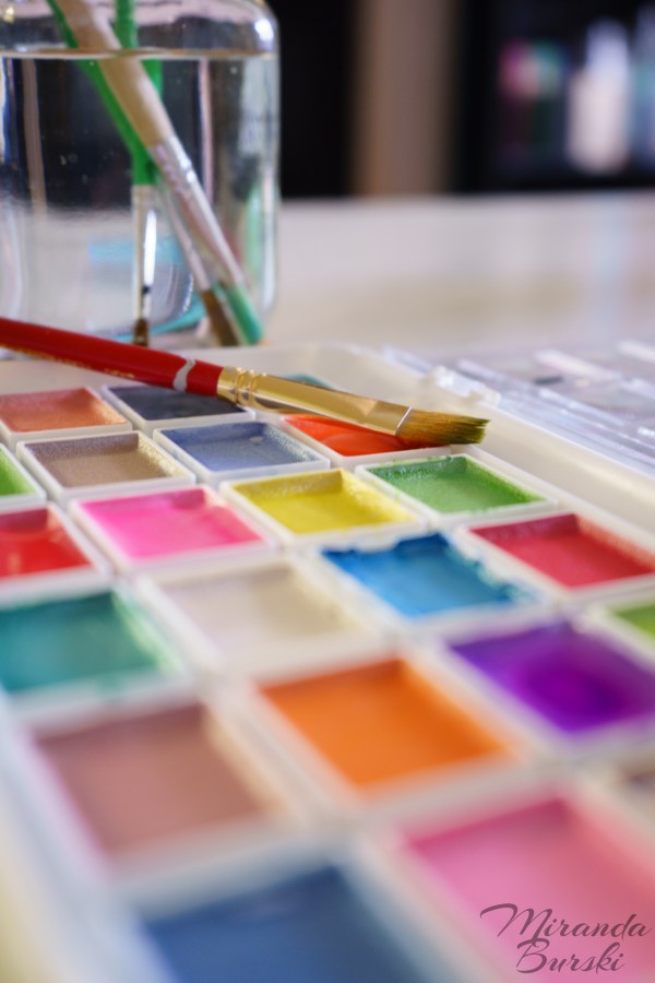 A paint brush sitting on a watercolour palette, with more brushes and water in the background.