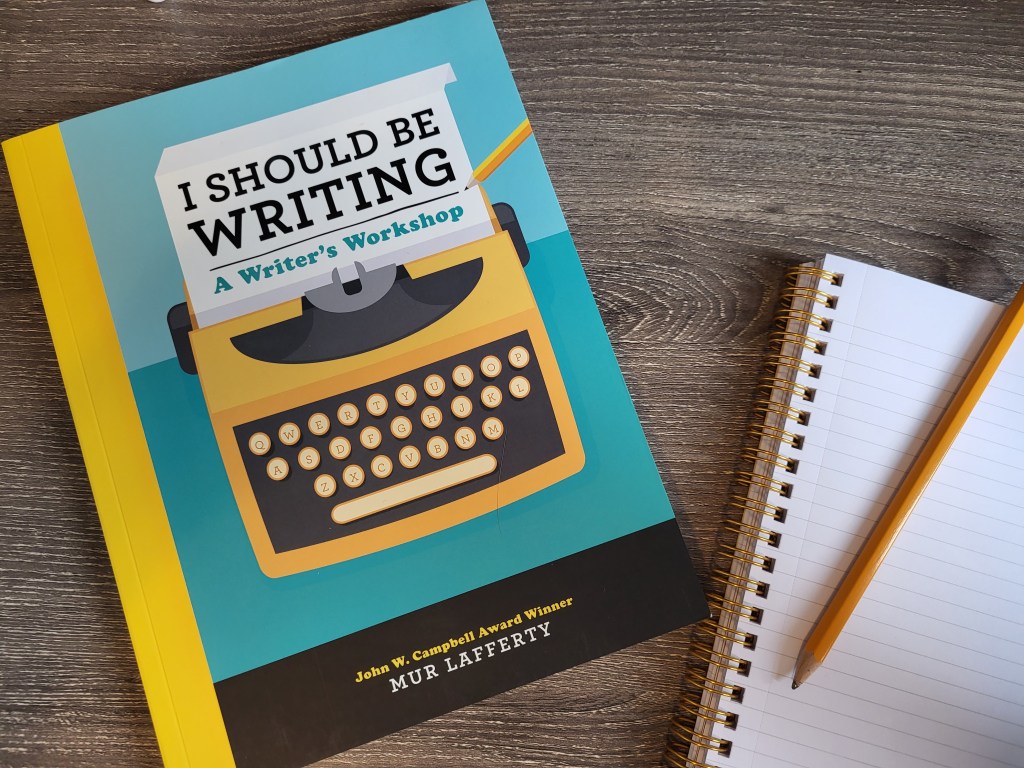 A book (I Should Be Writing by Mur Lafferty) set beside a notebook and pencil.